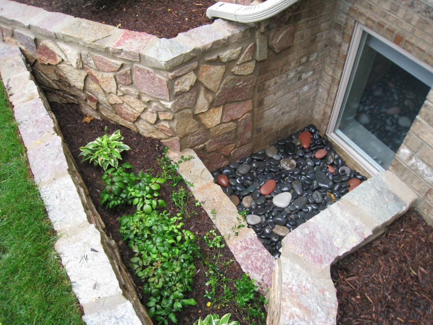 A window well opening into a planted bed, edged with dark stone